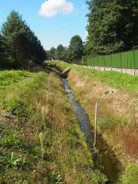 Wąski ciek o brzegach wyłożonych betonowymi ażurowymi płytami. Wzdłuż jednego z brzegów ciągnie się metalowe ogrodzenie