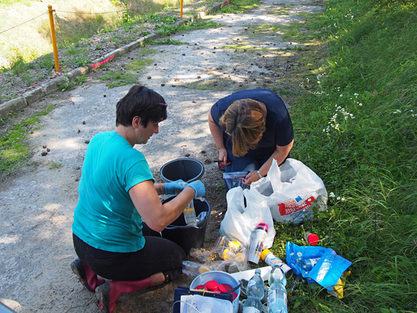 Dwie kobiety klęczą na ścieżce. Jedna z nich pisze markerem po plastikowym worku. Obok stoją wiadra i leżą butelki