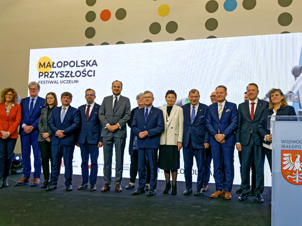 Grupa elegancko ubranych kobiet i mężczyzn stoi w szeregu na tle białego ekranu wyświetlającego logo festiwalu