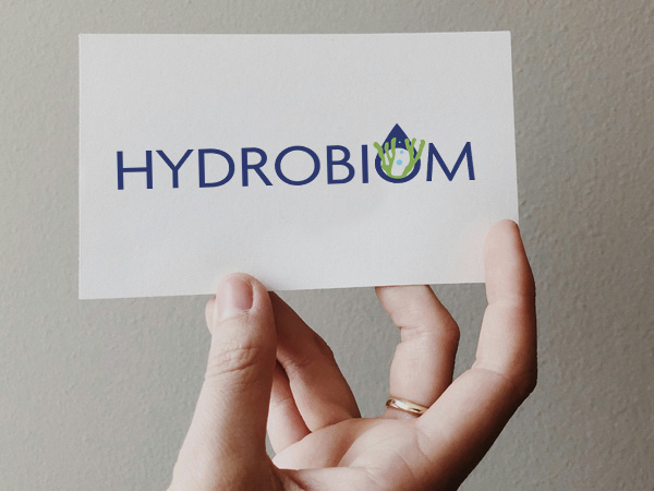 Kobieca dłoń trzyma kartkę z logo zawierającym granatowy napis Hydrobiom i rysunek glonu